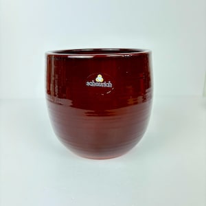 May include: A glossy, deep burgundy ceramic planter with a rounded shape. The planter has horizontal ridges and a small, white sticker with the brand name "scheurich". This decorative pot is suitable for indoor or outdoor use.
