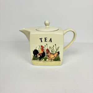 May include: Cream-colored ceramic teapot with a lid and handle. The front of the teapot features a painted scene of a rooster, hen, and the word "TEA". The teapot has a square shape with rounded edges.