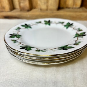 May include: Four white ceramic plates with gold rims, stacked. Each plate has a green and white leaf pattern around the edge. The plates are stacked on a white surface.