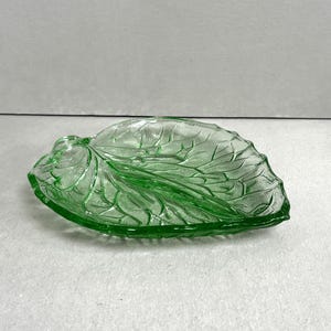 May include: A green glass leaf-shaped dish. The dish features detailed leaf vein patterns and a slightly textured surface. The colour is a vibrant, transparent green, and the dish is resting on a light-coloured surface.