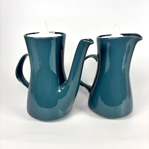 May include: Two teal ceramic serving jugs with white lids. One jug has a long spout, and the other has a shorter spout. Both have curved handles and a glossy finish. The jugs are likely for serving coffee or tea.