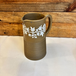May include: A brown ceramic pitcher with a white leaf design. The pitcher has a braided handle and a ribbed base. The white leaf design is around the top of the pitcher. The pitcher is on a white surface.
