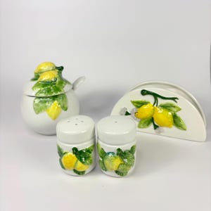 May include: A white ceramic kitchen set including a sugar bowl with a spoon, two salt and pepper shakers, and a napkin holder. Each item is decorated with yellow lemons and green leaves. The set is designed for the kitchen.