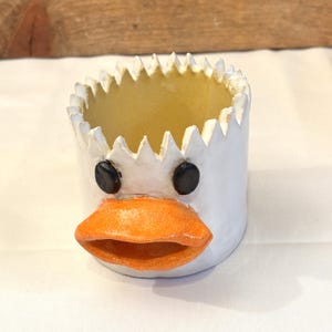 May include: A white ceramic duck-shaped planter with a jagged rim. The planter has black eyes and an orange beak. The interior is yellow. This whimsical container is suitable for small plants or decorative items.