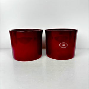 May include: Two matching, glossy, deep red ceramic planters. The planters have a cylindrical shape with a slightly flared rim. One planter has a small, white logo on the side. The planters are empty and ready for plants.