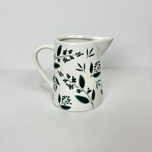 May include: A white ceramic pitcher with a curved handle and spout. The pitcher is decorated with a repeating pattern of dark green leaves and berries. The pitcher is suitable for serving beverages.
