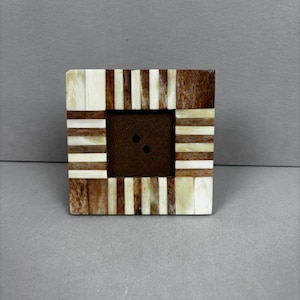 May include: Square photo frame with a mosaic design. The frame features alternating stripes of light beige and dark brown wood. The centre has a dark brown square with two small holes. The frame is set against a grey background.