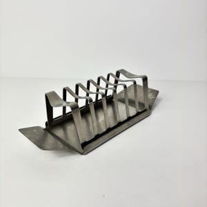 May include: A vintage silver-toned metal toast rack with six slots. The rack has a rectangular base and curved wire dividers. The design is simple and functional, suitable for holding multiple slices of toast.
