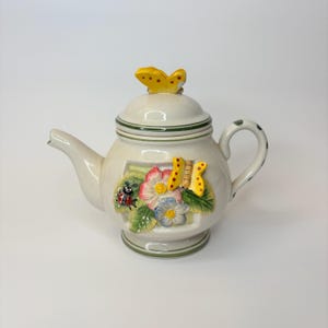 May include: Cream ceramic teapot with a lid and handle. The teapot is decorated with a yellow butterfly on the lid and a floral and insect design on the front. The handle has green polka dots.