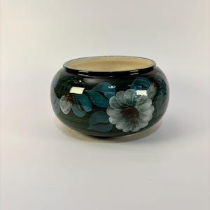 May include: A round, dark green ceramic bowl with a cream-colored interior. The exterior features hand-painted white and teal floral designs. The bowl has a glossy finish and is suitable for decorative or functional use.