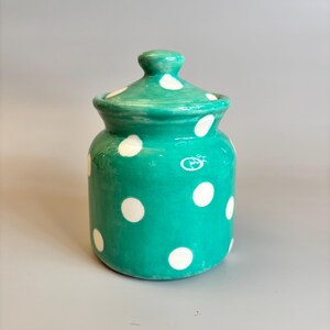 May include: A teal ceramic jar with a lid, decorated with large white polka dots. The jar has a rounded shape and a small knob on the lid. The jar is likely used for storage or decoration.
