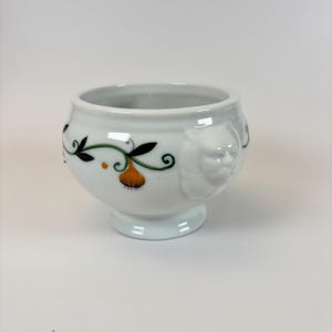 May include: White ceramic bowl with a pedestal base and a decorative lion's head on the side. The bowl features a painted design of orange fruit and green vines with black accents. The bowl is round with a wide opening.