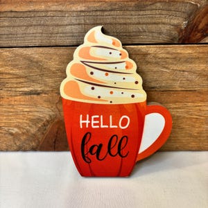 May include: Wooden fall-themed decoration shaped like a coffee mug. The mug is red with the words "HELLO fall" in white and black script. The top of the mug is topped with a swirl of cream-coloured whipped cream with brown and orange accents.