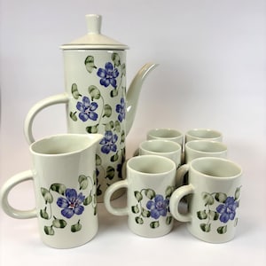 May include: A ceramic coffee set featuring a tall coffee pot with a lid, a creamer, and six mugs. The set is decorated with blue floral patterns and green leaves on a white background. The coffee pot measures approximately 25 cm tall.