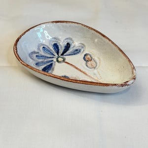 May include: Handmade ceramic dish with a white base, featuring a blue floral design and a brown rim. The dish has an oval shape and is suitable for serving small portions of food or as a decorative item.