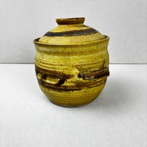 May include: A handmade ceramic jar with a lid. The jar is a warm yellow colour with dark brown horizontal stripes and accents. The lid has a small knob and matching stripes. The jar has a handle on the side.