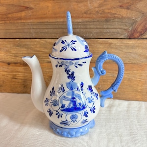 May include: A white and blue ceramic teapot with a windmill design. The teapot features a curved spout, a decorative handle, and a lid with a tall, curved handle. The design includes floral and windmill motifs, with blue accents on a white background.