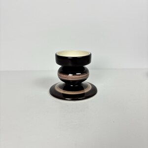 May include: A ceramic candle holder with a dark brown exterior and a cream-coloured interior. The holder has a circular base, a spherical midsection, and a cylindrical top. The design features concentric rings of varying shades of brown.