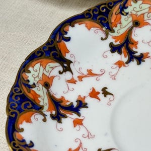 May include: A white porcelain saucer with a blue and gold rim. The saucer is decorated with a floral pattern in shades of orange, red, and green.