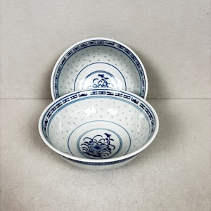 May include: Two white porcelain bowls with blue floral and geometric patterns. The bowls feature a delicate, perforated design and a blue floral motif in the centre. The rims are adorned with a blue geometric border.