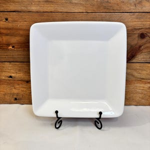May include: A square, white ceramic plate with rounded corners. The plate is displayed on a black metal stand with decorative scrollwork. The background is a wooden surface.
