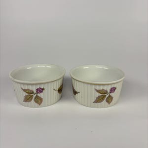 May include: Two white ceramic ramekins with vertical ribbed sides. Each ramekin features a floral design with a cluster of purple berries and green and brown leaves. The ramekins are ideal for baking and serving individual portions.