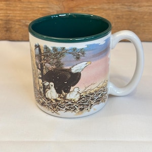 May include: White ceramic mug with a green interior rim and handle. The mug features a detailed illustration of a bald eagle with two chicks in a nest, set against a backdrop of a tree and a pink and blue sky. A nature-themed mug.
