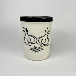 May include: A ceramic kitchen utensil holder with a cream-colored base and a black rim. The holder features a black illustration of two garlic bulbs and a cross-hatch design. The design is simple and elegant.