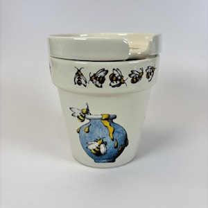 May include: A cream-coloured ceramic honey pot with a lid. The pot features a blue honey jar graphic with yellow honey dripping down, and cartoon bees. The rim of the pot has a repeating pattern of bees.