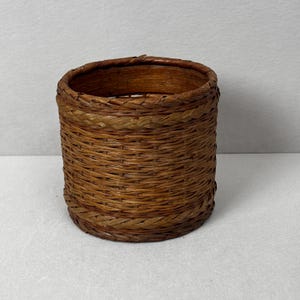May include: A cylindrical woven basket in natural brown tones. The basket is constructed from tightly woven reeds or similar material, featuring a braided trim at the top and bottom. The empty basket rests on a neutral surface.