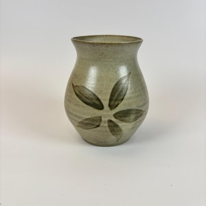 May include: A handcrafted ceramic vase in a muted green shade, decorated with a dark green floral motif. The vase has a rounded form and a gently flared rim. The design features six stylized leaves in a circular arrangement. Suitable for displaying flowers, it is a decorative item.