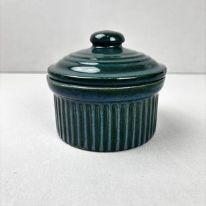 May include: A teal ceramic ramekin with a matching lid. The ramekin has a ribbed exterior and a rounded lid with a small knob. The colour is a deep teal with hints of blue, creating a classic look. Perfect for baking and serving individual portions.