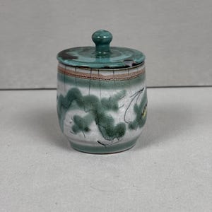 May include: A small, ceramic jar with a lid. The jar is cylindrical with a white base and green and brown accents. The lid and knob are a solid teal color. The jar has a hand-painted design of green foliage.