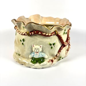 May include: A ceramic Christmas-themed container shaped like a bag, featuring a white cat figurine wearing a blue shirt and green trousers. The bag is cream-coloured with red and gold accents, holly leaves, and a red rope design.