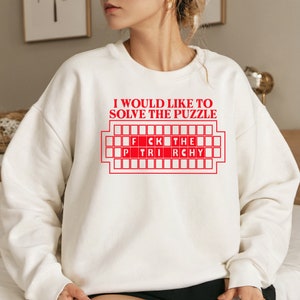 May include: White sweatshirt with red text that reads "I would like to solve the puzzle" and a red graphic of a game board with the words "FCK THE" and "P TRI RCHY" on it.