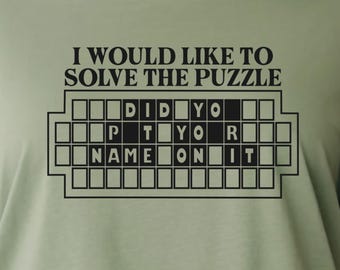 Pode incluir: Camiseta verde sálvia com um gráfico preto. O gráfico apresenta a frase "I WOULD LIKE TO SOLVE THE PUZZLE" acima de um tabuleiro de jogo com as palavras "DID YOU PUT YOUR NAME ON IT". Um colar de ouro é visível no topo.