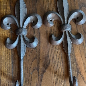 May include: Two cast iron fleur-de-lis wall hooks with a dark brown, rustic finish. Each hook features the classic three-petal design and a curved hook at the bottom. The hooks are designed for wall mounting and add a touch of vintage charm.
