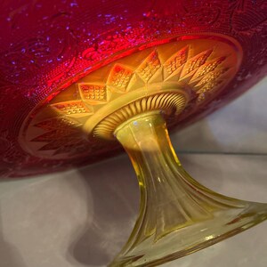 May include: A vintage, iridescent glass pedestal cake stand. The bowl is a deep red with etched floral patterns, transitioning to a golden-yellow base and stem. The stem has a flared base and a detailed, textured design.