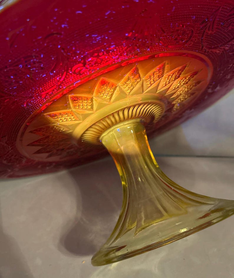 May include: A vintage, iridescent glass pedestal cake stand. The bowl is a deep red with etched floral patterns, transitioning to a golden-yellow base and stem. The stem has a flared base and a detailed, textured design.