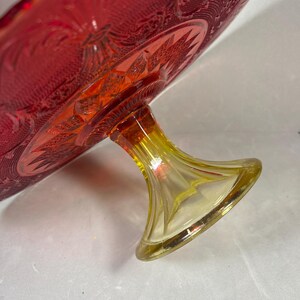 May include: A vintage red glass cake stand with a yellow base. The cake stand has a detailed, embossed pattern on the plate and a flared base. The glass has a rich, vibrant color.