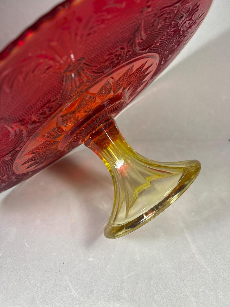 May include: A vintage red glass cake stand with a yellow base. The cake stand has a detailed, embossed pattern on the plate and a flared base. The glass has a rich, vibrant color.