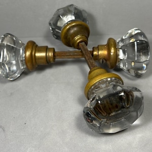 May include: Four vintage glass door knobs with brass hardware. The knobs are clear, faceted, and have a classic design. The brass components show signs of age and wear, adding to their antique charm. These knobs are suitable for restoration projects.