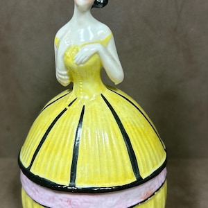 May include: A vintage porcelain figurine of a woman in a yellow dress with black stripes and a pink ribbon. The figurine is a trinket box with a hinged lid.
