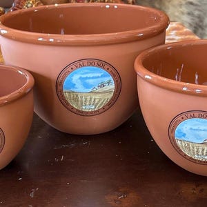 Puede incluir: Tres cuencos de terracota de diferentes tamaños. Cada cuenco tiene una forma redondeada con un exterior liso y sin esmaltar y un interior de color más claro. Cada cuenco tiene una etiqueta circular con un paisaje.
