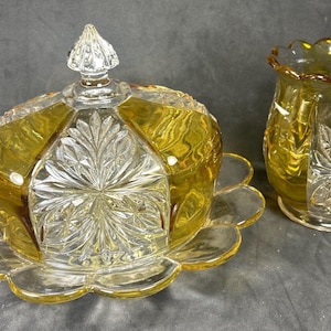 May include: A butter dish with a clear and amber glass dome lid featuring a pointed finial and a scalloped base, accompanied by a matching amber and clear glass vase. Both pieces display a pressed glass pattern with starburst motifs.