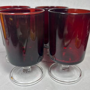 Puede incluir: Cuatro copas de vino con pie de color rojo rubí. Las copas tienen una base circular transparente y un cuenco cilíndrico. El borde de cada copa es de color rojo intenso, con el color desvaneciéndose hacia abajo. Las copas están juntas.