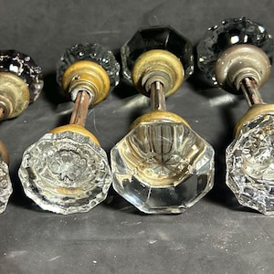 May include: Four antique glass door knobs with brass hardware. The knobs have faceted, clear glass bases and dark, possibly black, tops. The brass hardware shows signs of age and wear, adding to the vintage aesthetic. These knobs are likely from an older home.