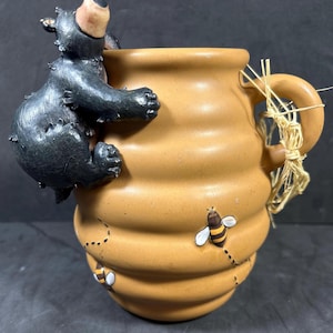 Puede incluir: Una taza de cerámica con forma de tarro de miel con un oso negro trepando por el lateral. La taza es de color marrón claro con un diseño acanalado y presenta abejas pintadas. El asa está adornada con paja.