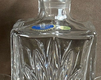May include: A clear, faceted crystal decanter with a matching stopper. The decanter has a square base and a detailed, textured design. The stopper is a large, round crystal ball. A classic barware item.