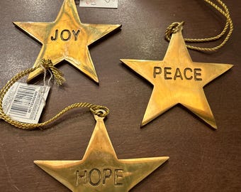 May include: Three gold-coloured star-shaped Christmas ornaments. Each star has a word engraved: "JOY", "PEACE", and "HOPE". They have gold string hangers and are on a brown surface.